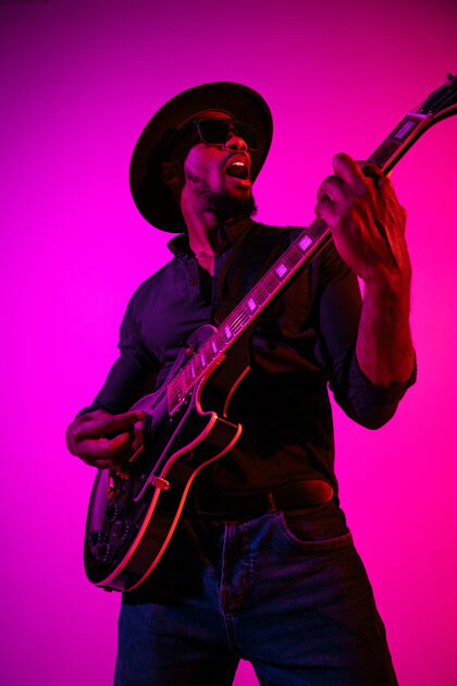娱乐年轻的非洲裔美国音乐家在霓虹灯下像摇滚明星一样在渐变的紫粉色背景上弹吉他时尚站立酒廊