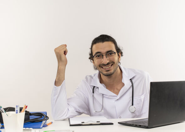 笔记本电脑微笑的年轻男医生戴着医用眼镜 穿着带听诊器的医用长袍手势工具男性