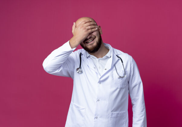 男性面带微笑的年轻男医生穿着医用长袍 戴着听诊器 用手遮住眼睛 隔离在粉红色的皮肤上医生听诊器微笑