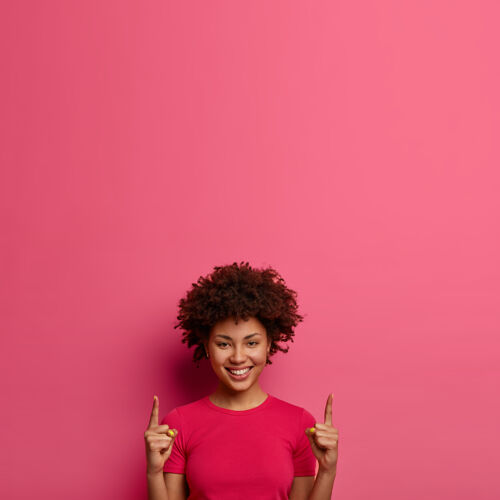 欢呼快乐的卷发女青年的垂直镜头显示在上面 显示头上的空白 高兴地微笑 显示白色的牙齿 显示向上的东西 孤立在粉红色的墙上空白微笑手势