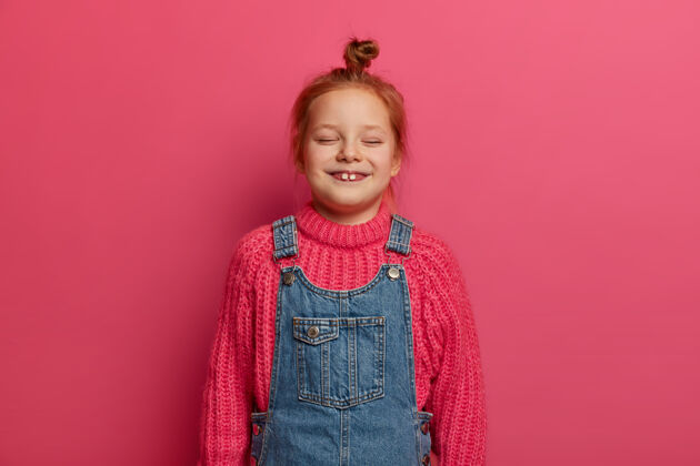 编织五岁的小女孩 姜黄色的发髻和两颗突出的牙齿 穿着暖和的针织毛衣和牛仔服 欢笑着 闭着眼睛 看着有趣的卡通 孤立在粉红色的墙上狐狸模特室内