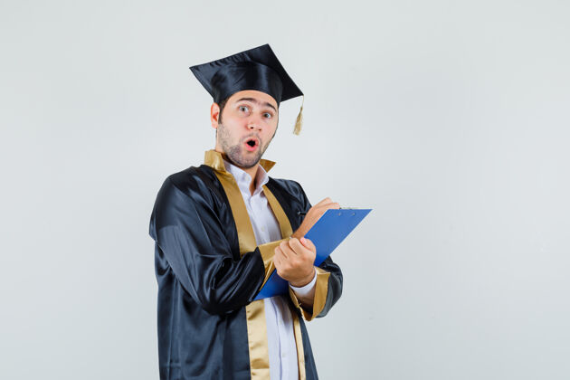 年轻年轻人穿着毕业制服在剪贴板上做笔记 看上去很惊讶前视图英俊成人成功