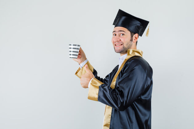 拇指年轻人拿着一杯饮料 穿着毕业制服竖起大拇指 看上去很高兴聪明休闲自信