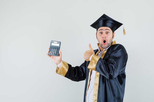 肖像年轻人拿着计算器 穿着毕业制服竖起大拇指 看上去很惊讶前视图英俊毕业计算器