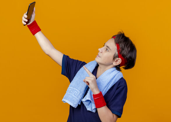 运动年轻英俊的运动男孩戴着头带和护腕 脖子上戴着牙套和毛巾 自拍时指着橘色墙上的手机脸脖子周围