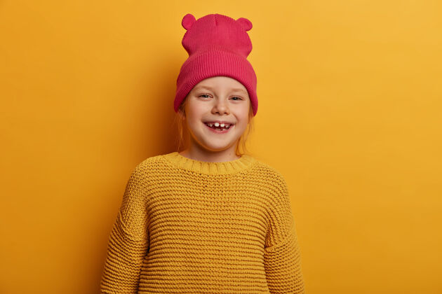 感觉表情满意的快乐小女孩表情直率 微笑积极 表达真挚的情感 心情乐观 戴着带耳朵的粉色帽子和编织的黄色毛衣 在室内摆姿势乐观室内孩子