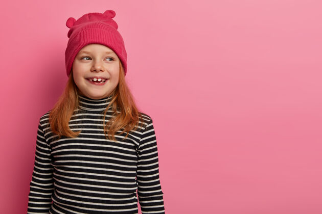 帽子红发积极的女孩戴着帽子和条纹圆领衫 看向一边 注意到一些有趣的事情 记得和父母度过了美好的时光 在粉彩玫瑰色的墙上摆姿势 有可爱的表情女儿感觉时尚
