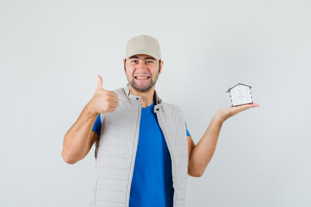 站年轻人拿着房子模型 展示拇指在t恤衫 夹克衫 帽子和期待喜悦前视图房子胡须夹克