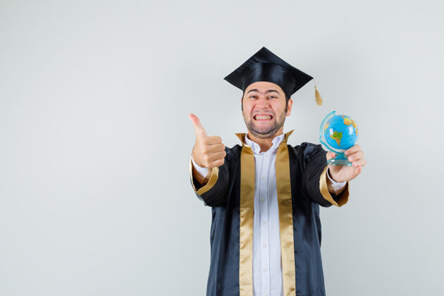 地球仪年轻人拿着学校的地球仪 穿着研究生制服竖起大拇指 看上去很幸运 正前方视图年轻男人肖像