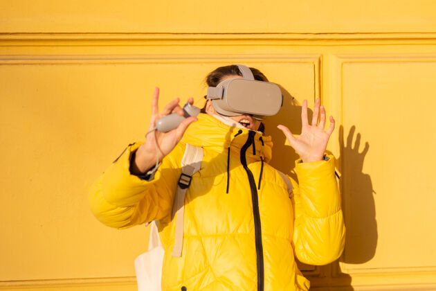 虚拟现实游戏一个戴着虚拟现实眼镜的女人的肖像 在冬天的阳光下 穿着暖和的衣服 靠在黄色的墙上不高兴视频设备