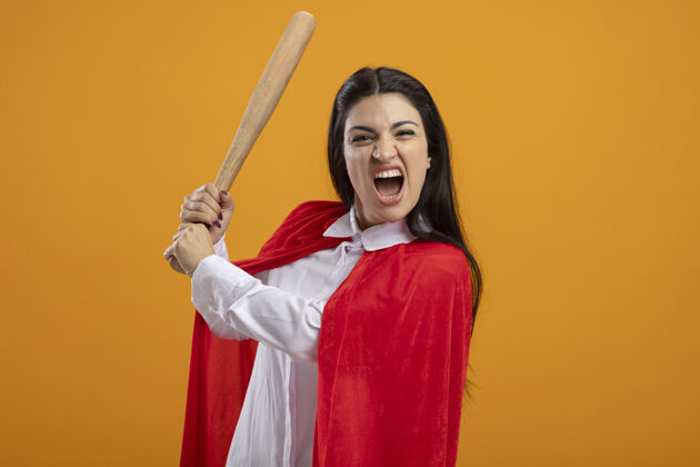 愤怒愤怒的年轻女超人拿着棒球棒看着橙色墙上孤立的前方衣服人脸