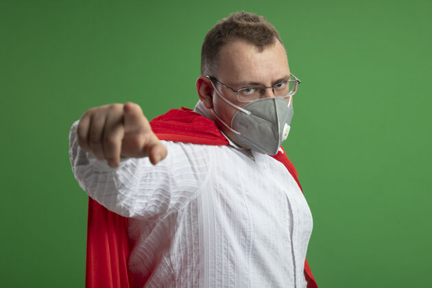 帽子自信的成年超级英雄身穿红色斗篷 戴着眼镜和防护面具 站在侧视图中 看着并指着隔离在绿色墙上的前方立场脸指向