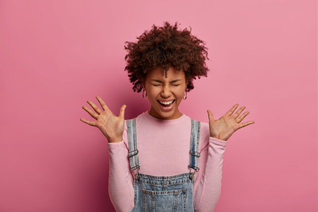 休闲活泼积极的非洲裔美国年轻女子抬起手掌 为听到惊人的消息而发狂 笑出声来 穿着高领毛衣和萨拉芬 在粉红色的墙上摆姿势快乐的情感概念疯狂萨拉凡积极
