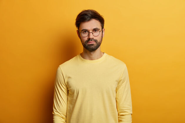 站立照片中严肃的男人有深色的鬃毛 戴着圆眼镜和黄色的套头衫 直视 在室内摆姿势 与人随意交谈单色面部表情概念男人成人脸