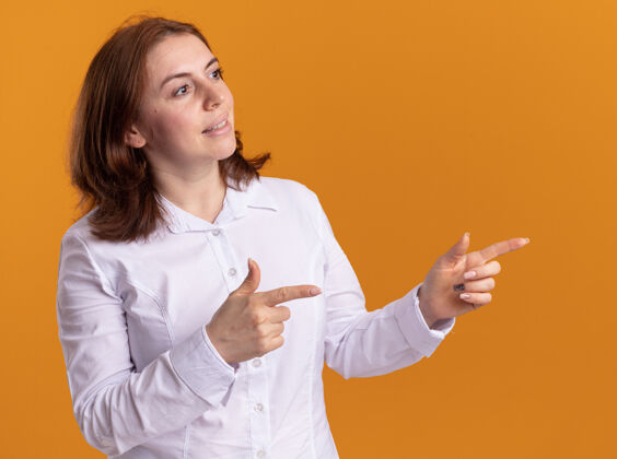 衬衫身穿白衬衫的年轻女子自信地微笑着 一边看一边用食指指着站在橙色墙上的一边手指微笑侧身