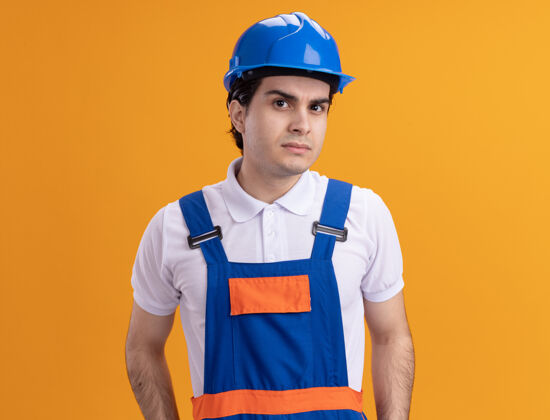 站着身穿施工制服 头戴安全帽的年轻建筑工人站在橙色的墙上 带着怀疑的表情看着前方年轻表情头盔