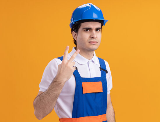 脸年轻的建筑工人穿着建筑制服 戴着安全帽 严肃地看着前面 二号站在橙色的墙上表演建筑严肃