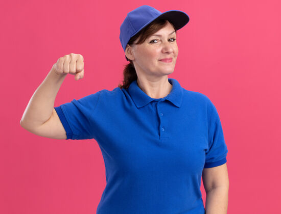 制服身穿蓝色制服 头戴鸭舌帽的中年女送货员站在粉红色的墙上 看着前面紧握的拳头 微笑着自信地站着显示帽子握紧