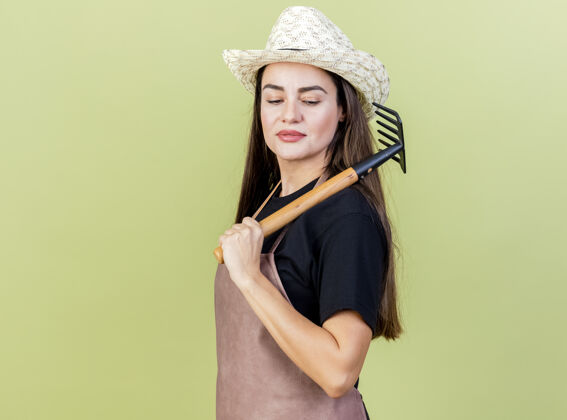 园艺自信美丽的园丁女孩穿着制服戴着园艺帽把耙子放在肩膀上孤立在橄榄绿的背景上橄榄帽子耙子