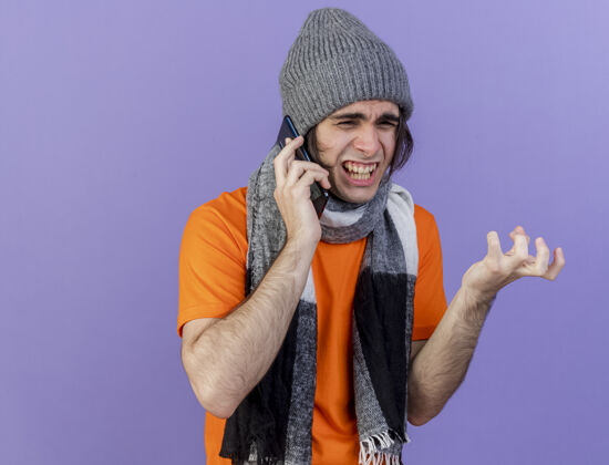 说话愤怒的年轻病夫戴着冬天的帽子 戴着围巾 在紫色的背景下讲电话年轻电话愤怒