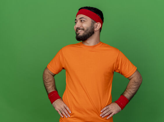 腕带面带微笑的年轻运动型男子 戴着头带和腕带 手放在臀部 与绿色背景隔离壁板年轻运动