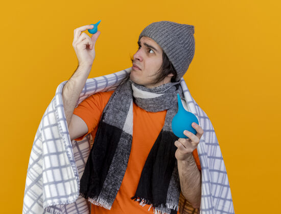 帽子困惑的年轻病人戴着冬天的帽子 围巾裹着格子布 抬头望着隔离在橙色背景上的灌肠剂灌肠养育包扎