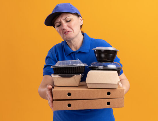 披萨身穿蓝色制服 头戴鸭舌帽的中年女送货员站在橙色的墙上 手里拿着披萨盒和食品包 困惑地 不高兴地看着它们年龄送货站着