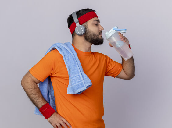运动站在侧视图中的年轻运动型男子戴着头带和腕带 戴着耳机 从水瓶里喝水 手放在臀部 肩上放着毛巾 隔离在白色背景上手立场瓶子