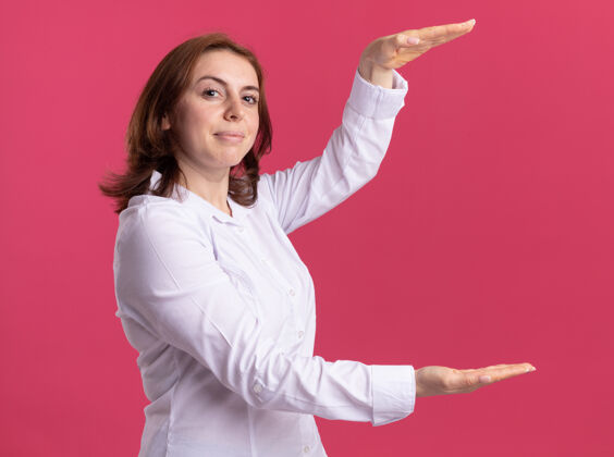 立场身穿白衬衫的年轻女子举手表示身材 测量符号微笑自信地站在粉红色的墙上女人手衬衫