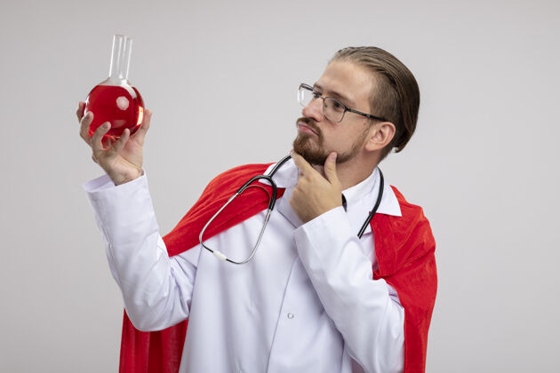 年轻年轻的超级英雄穿着医用长袍 戴着听诊器和眼镜 拿着化学玻璃瓶 里面装满了红色液体 手放在下巴上 隔离在白色背景上长袍思考瓶子