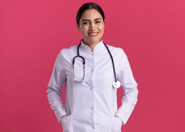 站着年轻的女医生穿着医用外套 手持听诊器 站在粉红色的墙上 面带微笑地看着前方年轻女人听诊器