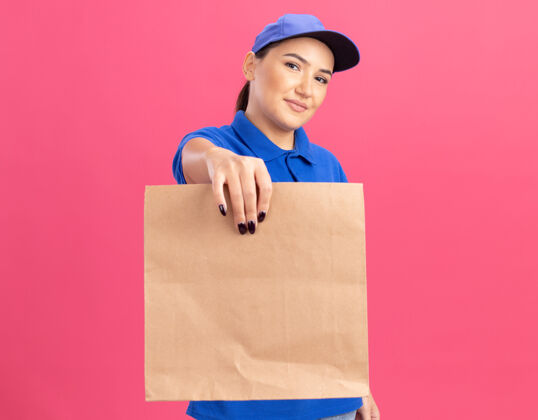 微笑身穿蓝色制服 头戴鸭舌帽的年轻女送货员拿着纸包望着前方 微笑着自信地站在粉色的墙上女人送货自信