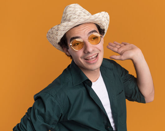 积极一个穿着绿色衬衫 戴着夏季帽子 戴着眼镜的年轻人站在橘色的墙上 高兴而积极地看着一边 举起手臂举起眼镜衬衫