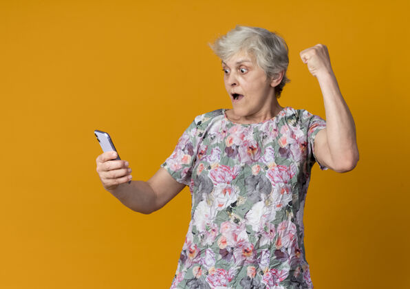 老的惊讶的老妇人举起拳头看着隔离在橙色墙上的电话老人起来女人