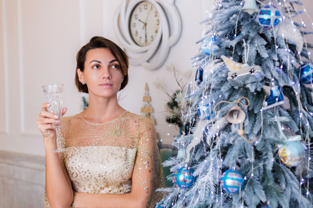 豪华一个穿着闪亮的金色圣诞晚礼服的女人在家里拿着一杯香槟在圣诞树旁优雅玻璃酒精