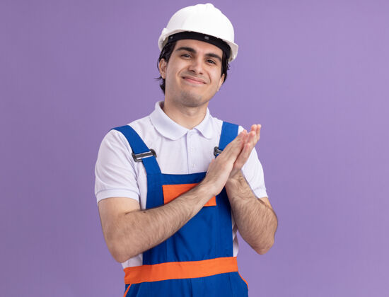建设者年轻的建筑工人穿着建筑制服 戴着安全帽 站在紫色的墙上 微笑着自信地鼓掌鼓掌安全建筑
