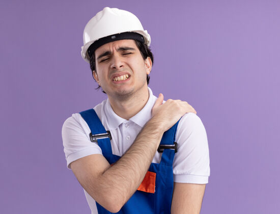 安全身穿施工制服 头戴安全帽的年轻建筑工人站在紫色的墙壁上 看上去身体不适 摸着肩膀感到疼痛触摸男人感觉