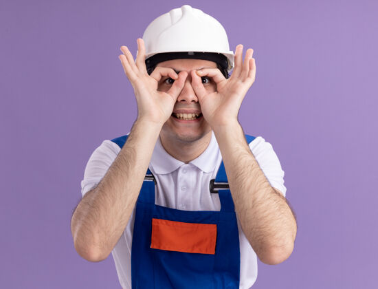 男人身穿建筑制服 戴安全帽的年轻建筑工人站在紫色的墙壁上 用手指做着双眼微笑的姿势注视着前方望远镜微笑站着