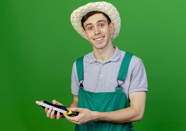 磁带微笑的年轻男园丁戴着园艺帽 用卷尺量着茄子帽子微笑花园