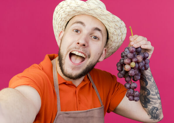 葡萄快乐的男园丁戴着园艺帽拿着葡萄假装拿着相机复制粉色喜悦