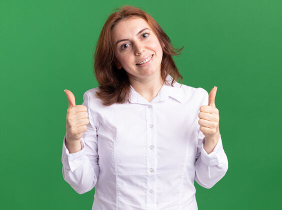 衬衫身穿白衬衫的快乐年轻女子站在绿色的墙上 面带微笑地看着前面 竖起大拇指脸站起来微笑