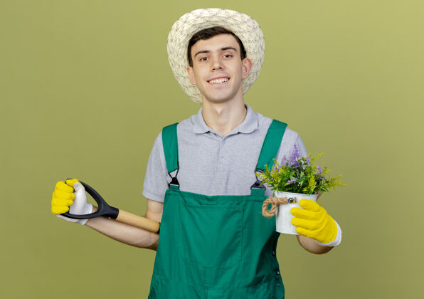 帽子微笑着的年轻男园丁戴着园艺帽和手套 手里拿着花盆里的花 背后拿着铲子持有花园橄榄