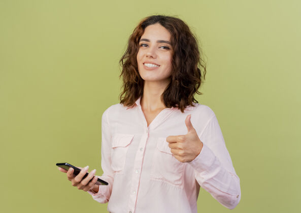 背景微笑的年轻白种女人拿着手机 竖起大拇指孤立地站在绿色背景上 留着复印空间女人抱着年轻