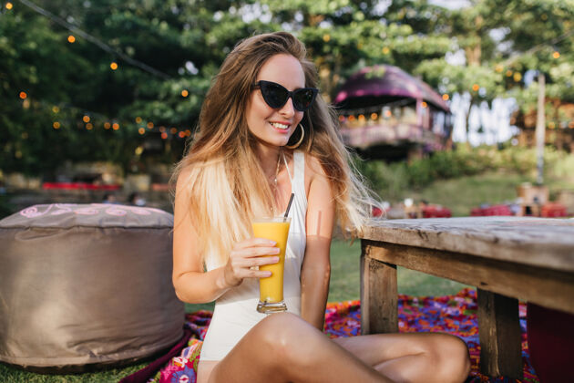 成人美丽的金发女人戴着太阳镜在公园里喝着橙色鸡尾酒乐趣冥想身体