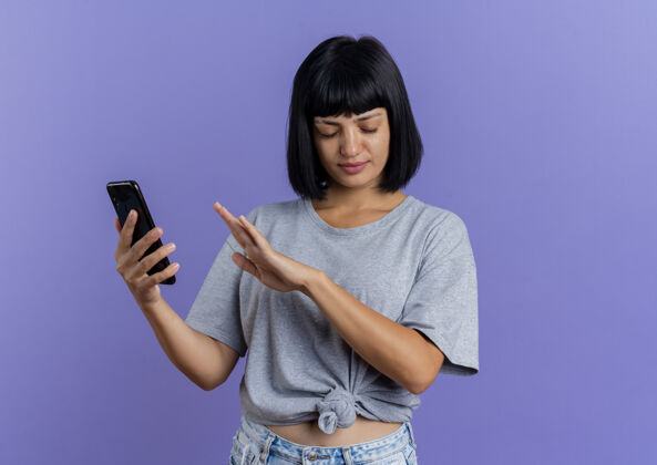 烦恼恼怒的年轻黑发白种女人拿着电话 手牵着屏幕前的紫色背景与复制空间隔离手女人电话