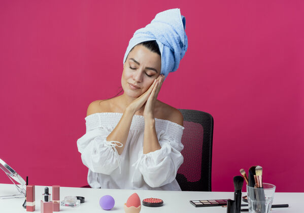 包困倦美丽的女孩裹着毛巾坐在桌旁 用化妆工具双手合十 脸贴在粉红色的墙上化妆粉色坐