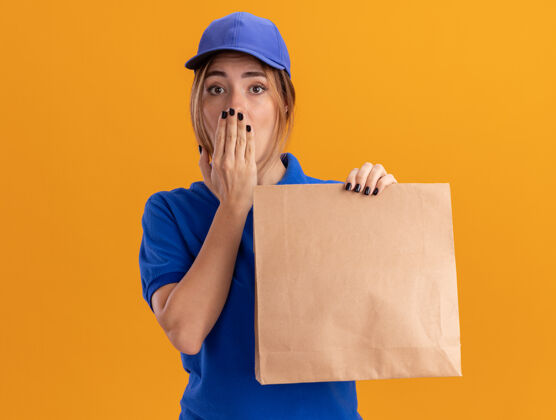 交货焦急的年轻漂亮的女送货员穿着制服 把手放在嘴巴上 手里拿着橘子色的纸包年轻包装手