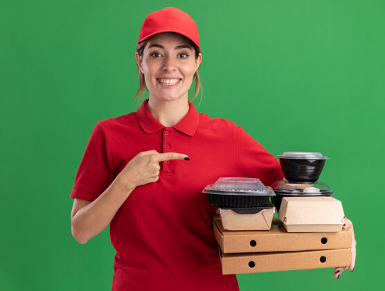姿势微笑着的年轻漂亮的女送货员穿着制服 指着绿色的比萨饼盒上的纸食品包装和容器漂亮送货成人