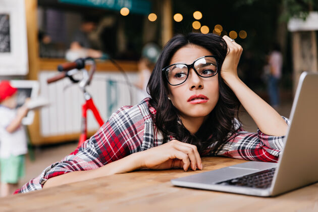 电脑沉思的黑发自由职业者坐在户外 望着别处疲惫的女留学生戴着眼镜用电脑的肖像户外女人青年