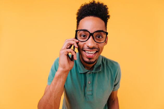 人有兴趣的戴眼镜的黑眼睛男人在打电话快乐的非洲男人穿着绿色衣服和智能手机摆姿势冷快乐沟通